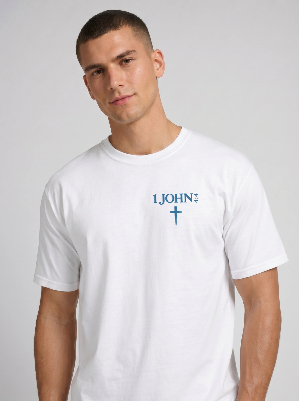 A man wearing a white garment-dyed t-shirt, viewed from the front. The shirt has a small chest print reading '1 JOHN 4:4' with a cross symbol beneath it.