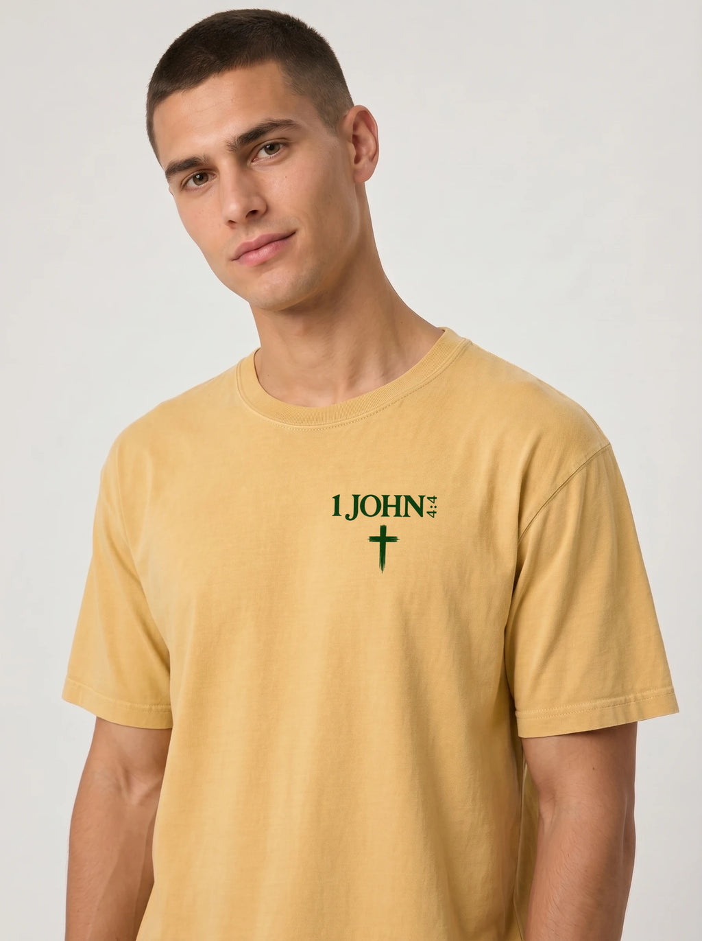Person wearing a faded yellow T-shirt with green “1 John 4:4” text and a cross printed on the chest.