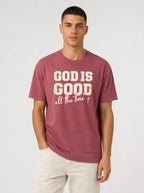 A man wearing a maroon crewneck t-shirt with a distressed white graphic that reads 'GOD IS GOOD all the time +', paired with off-white pants, against a light gray studio background.