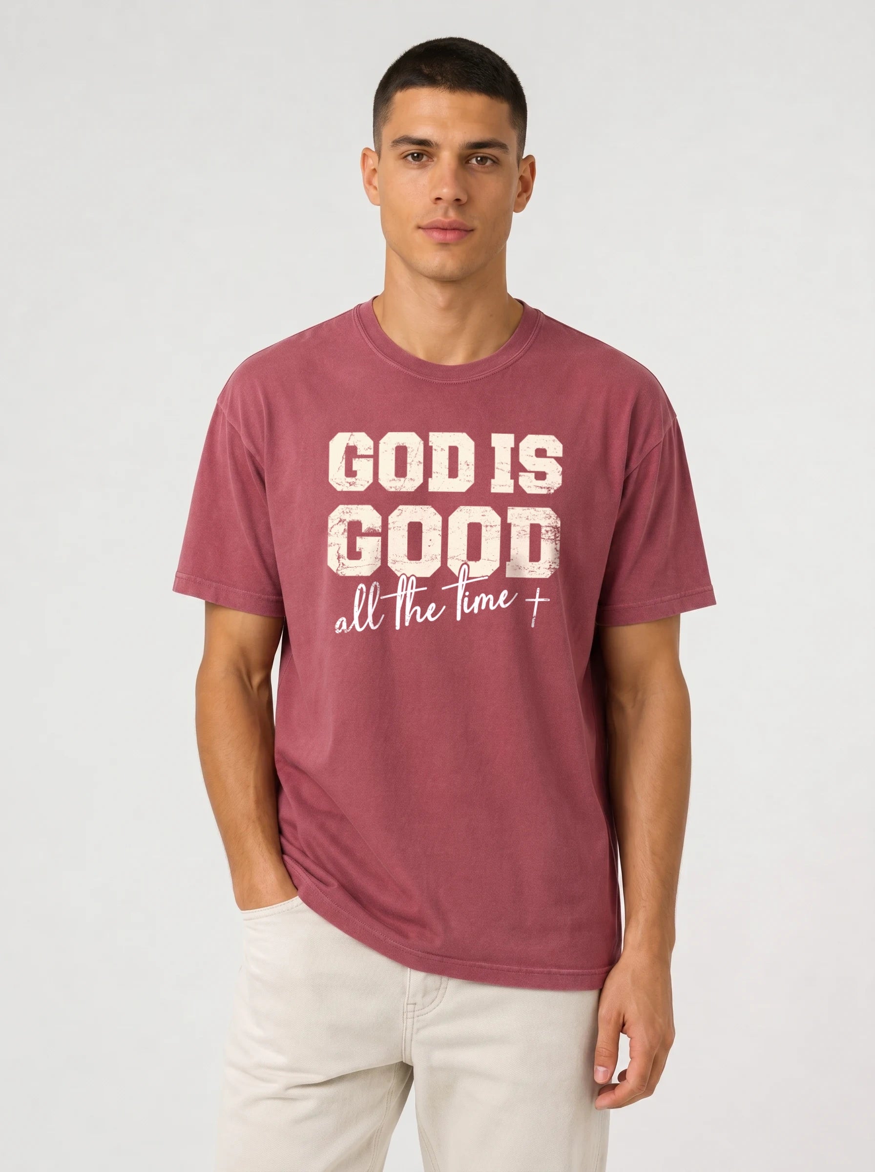 A man wearing a maroon crewneck t-shirt with a distressed white graphic that reads 'GOD IS GOOD all the time +', paired with off-white pants, against a light gray studio background.