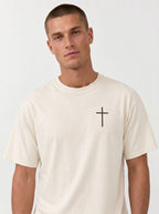 A man wearing a cream-colored crewneck t-shirt with a small black cross printed on the left chest, photographed against a light gray studio background.