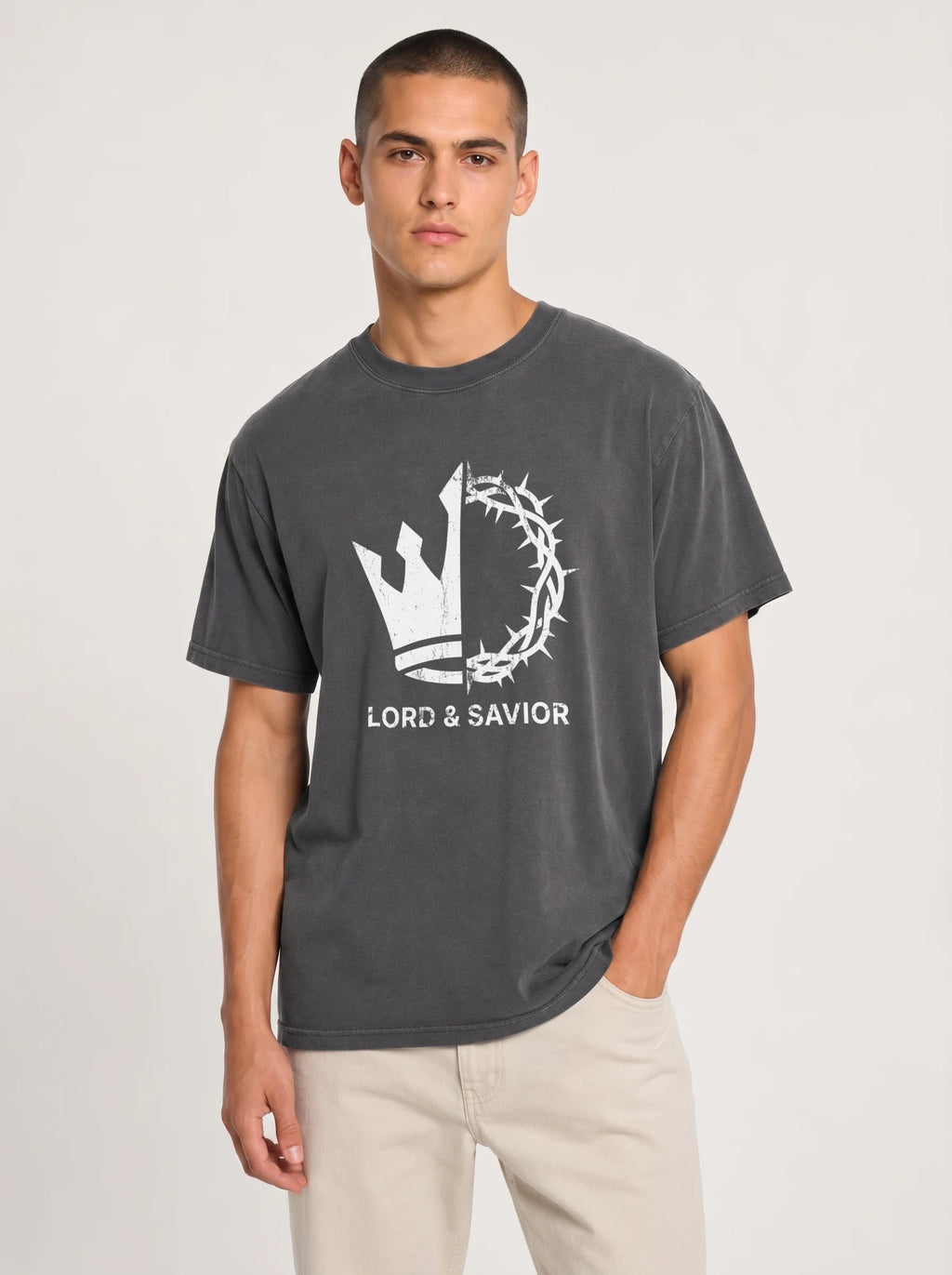 A man wearing a dark gray crewneck t-shirt with a white graphic of a crown of thorns and the text 'LORD & SAVIOR' underneath, paired with light beige pants, against a light gray studio background.