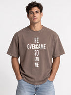Person wearing a faded brown T-shirt with large white text reading “HE OVERCAME SO CAN WE.”