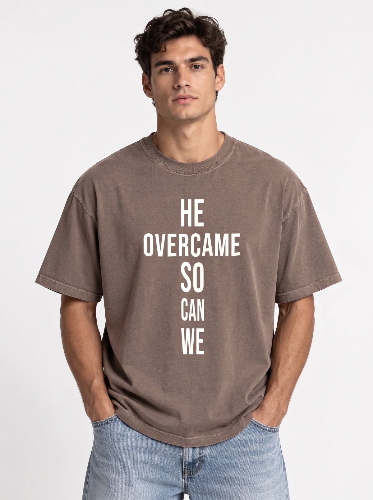 Person wearing a faded brown T-shirt with large white text reading “HE OVERCAME SO CAN WE.”