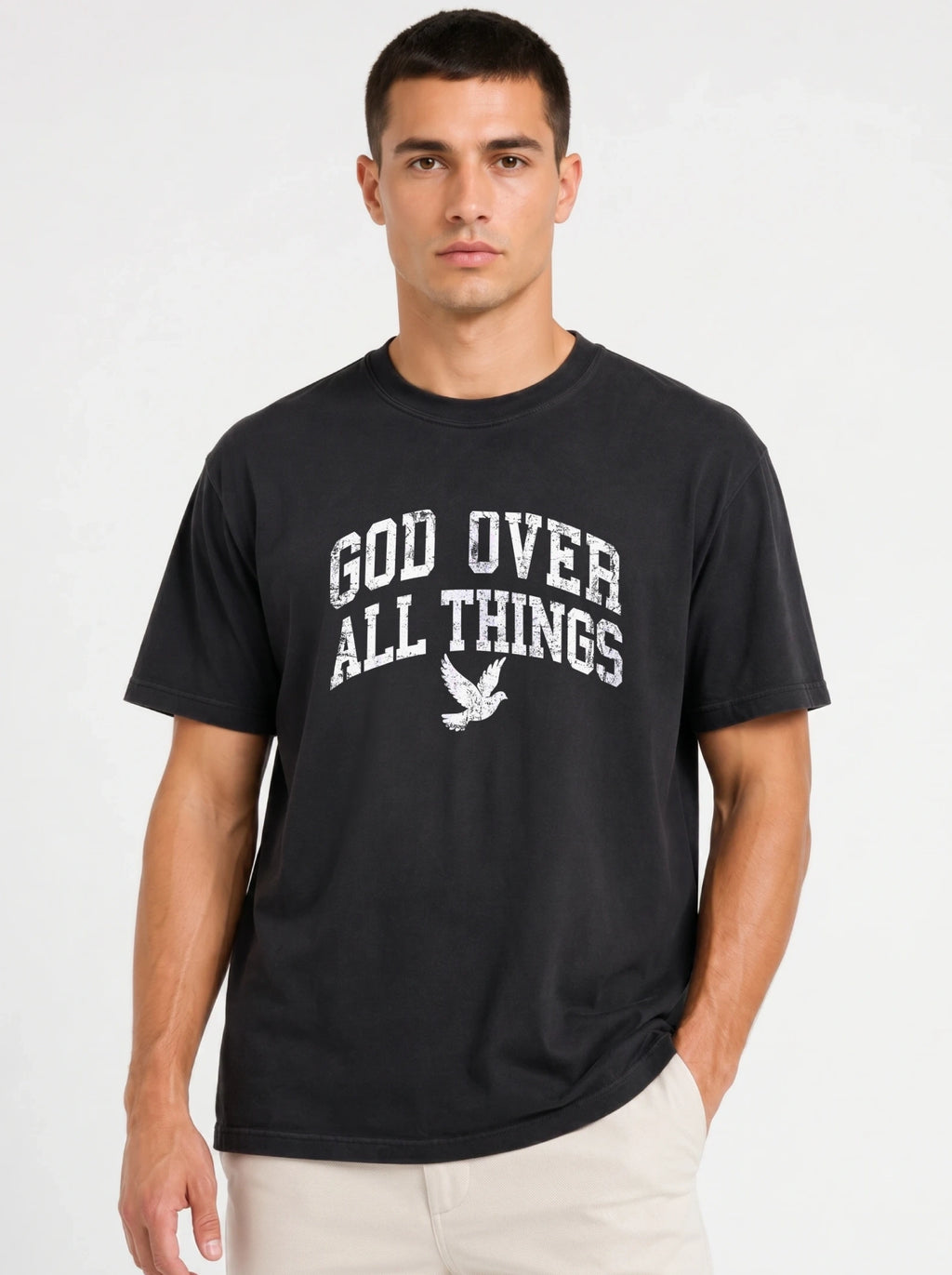 Medium front-facing shot of a male model wearing a solid black short-sleeve t-shirt with his hand in his pocket, displaying a distressed white "GOD OVER ALL THINGS" graphic and dove logo.