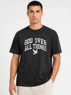 Medium front-facing shot of a male model wearing a solid black short-sleeve t-shirt with his hand in his pocket, displaying a distressed white "GOD OVER ALL THINGS" graphic and dove logo.