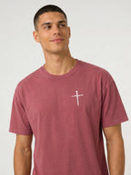 A man wearing a dusty rose short-sleeve t-shirt with a white cross graphic across the chest, set against a plain light gray studio background.