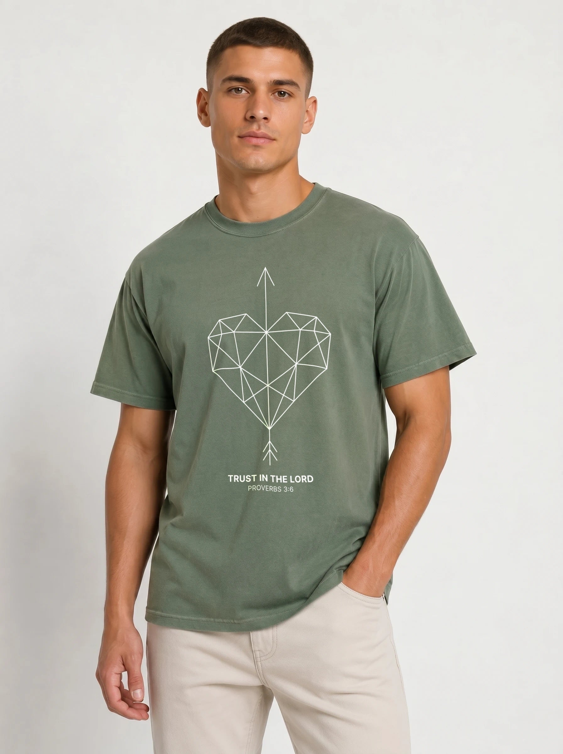 A man wearing an olive green crewneck t-shirt with a geometric heart pierced by an upward-pointing arrow, paired with the verse Proverbs 3:6: "Trust in the Lord.", paired with light beige pants, against a plain light gray background.