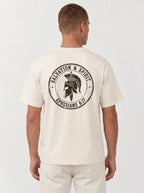A man viewed from the back wearing a cream-colored t-shirt with a large black circular graphic on the back that reads 'Salvation & Spirit Ephesians 6:17' around a helmet design, paired with cream pants against a light gray studio background.