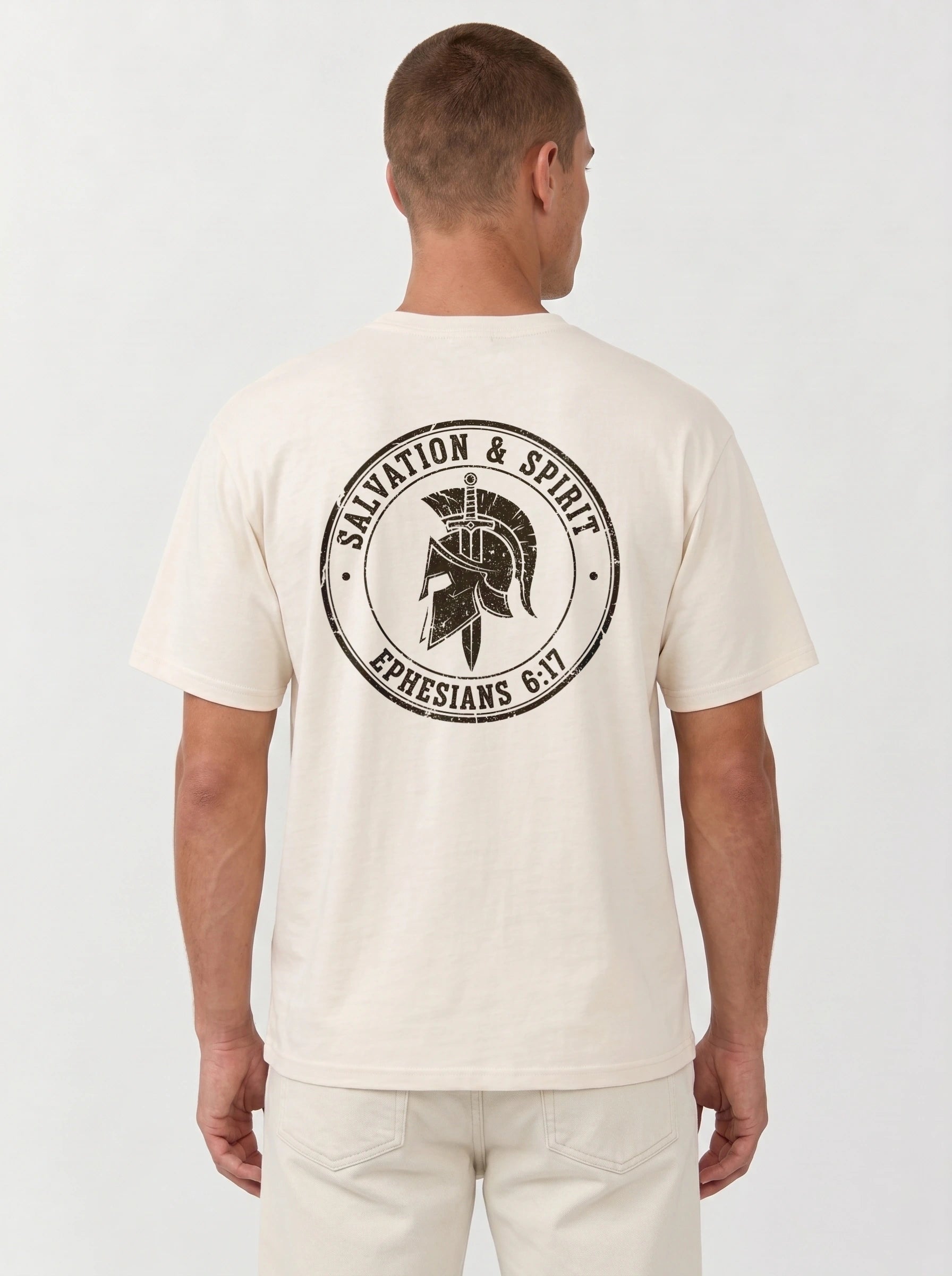 A man viewed from the back wearing a cream-colored t-shirt with a large black circular graphic on the back that reads 'Salvation & Spirit Ephesians 6:17' around a helmet design, paired with cream pants against a light gray studio background.