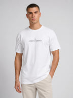 A man wearing a white short-sleeve crewneck t-shirt with the text 'JESUS SAVES' and a small cross printed on the chest, paired with beige pants, against a light gray studio background.