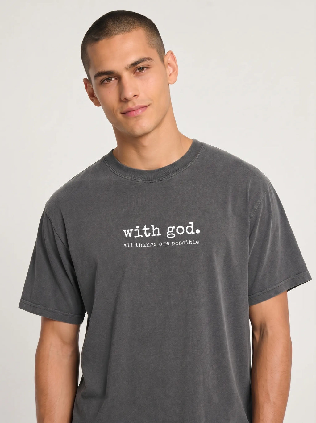 A man wearing a dark gray mineral-wash t-shirt with a vertical white line graphic and the text 'with god. all things are possible' printed on the chest, set against a plain light gray background.