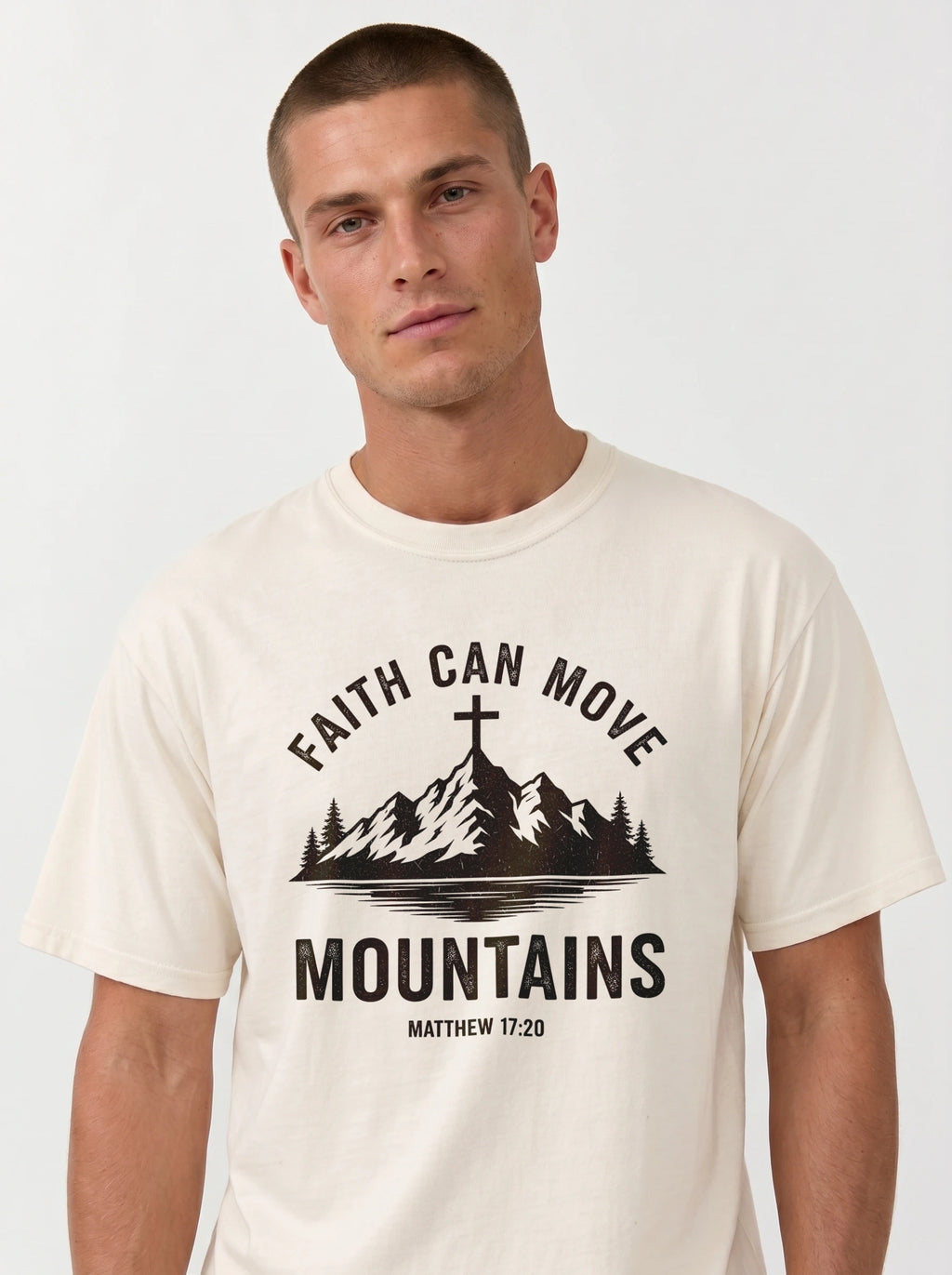 A man wearing a cream-colored t-shirt with a black graphic print that reads 'FAITH CAN MOVE MOUNTAINS' and 'MATTHEW 17:20' with a mountain and cross illustration.