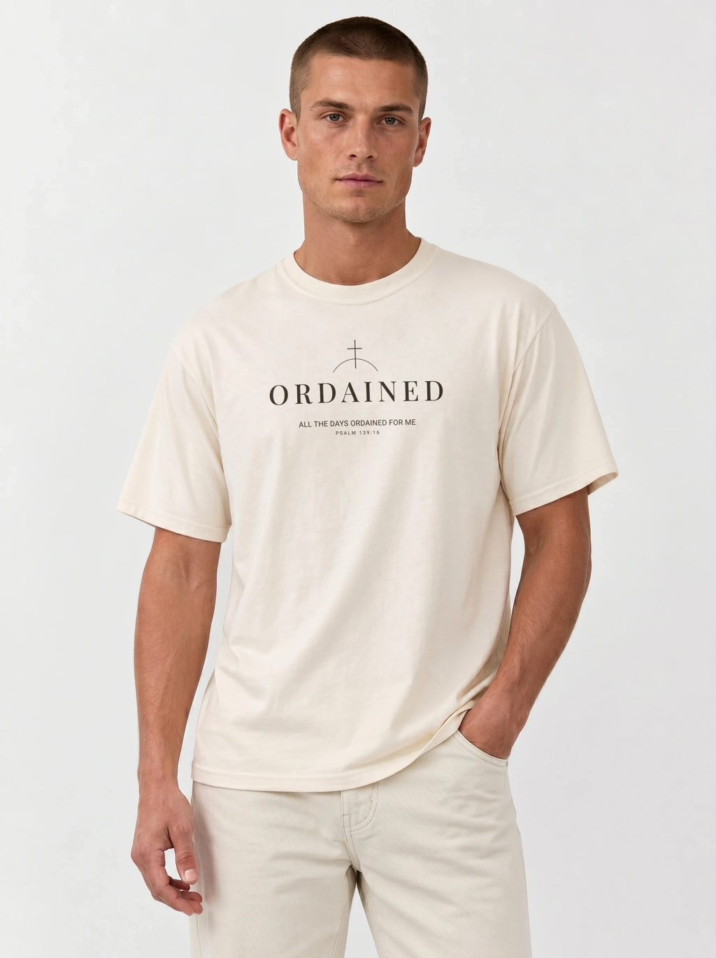 A man wearing a cream-colored crewneck t-shirt with 'ORDAINED' and a cross graphic printed on the chest, paired with matching cream pants, against a light gray studio background.