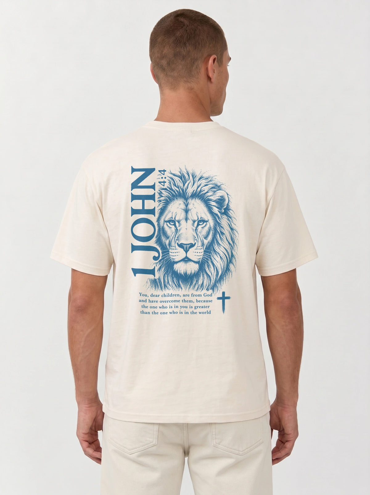 A man wearing a cream-colored garment-dyed t-shirt, viewed from the back at a closer angle. The shirt shows a large blue lion illustration with '1 JOHN 4:4' text, a Bible verse, and a small cross symbol.