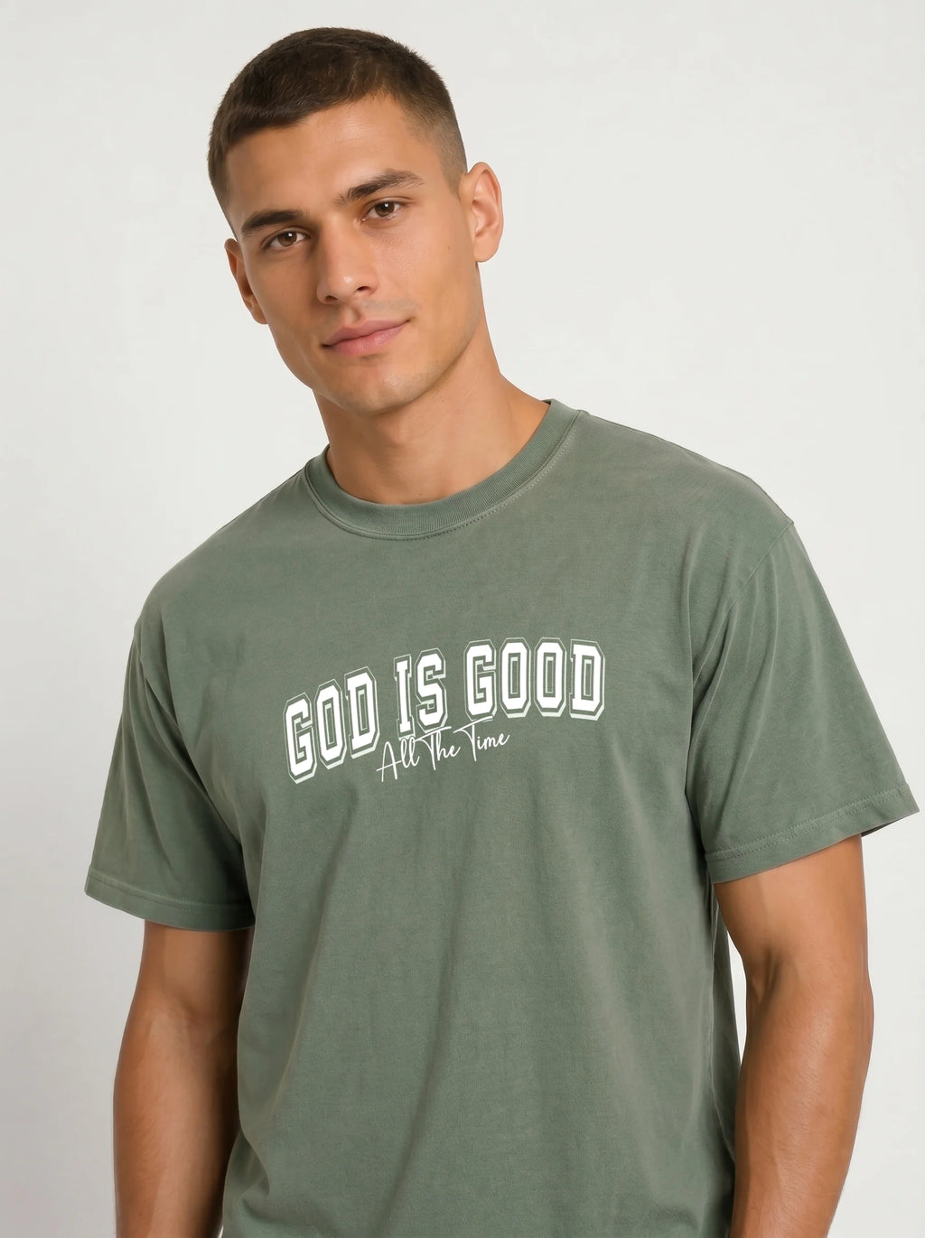 A man wearing a muted sage green t-shirt with the text 'GOD IS GOOD All The Time' printed in white across the chest, photographed against a light gray studio background.