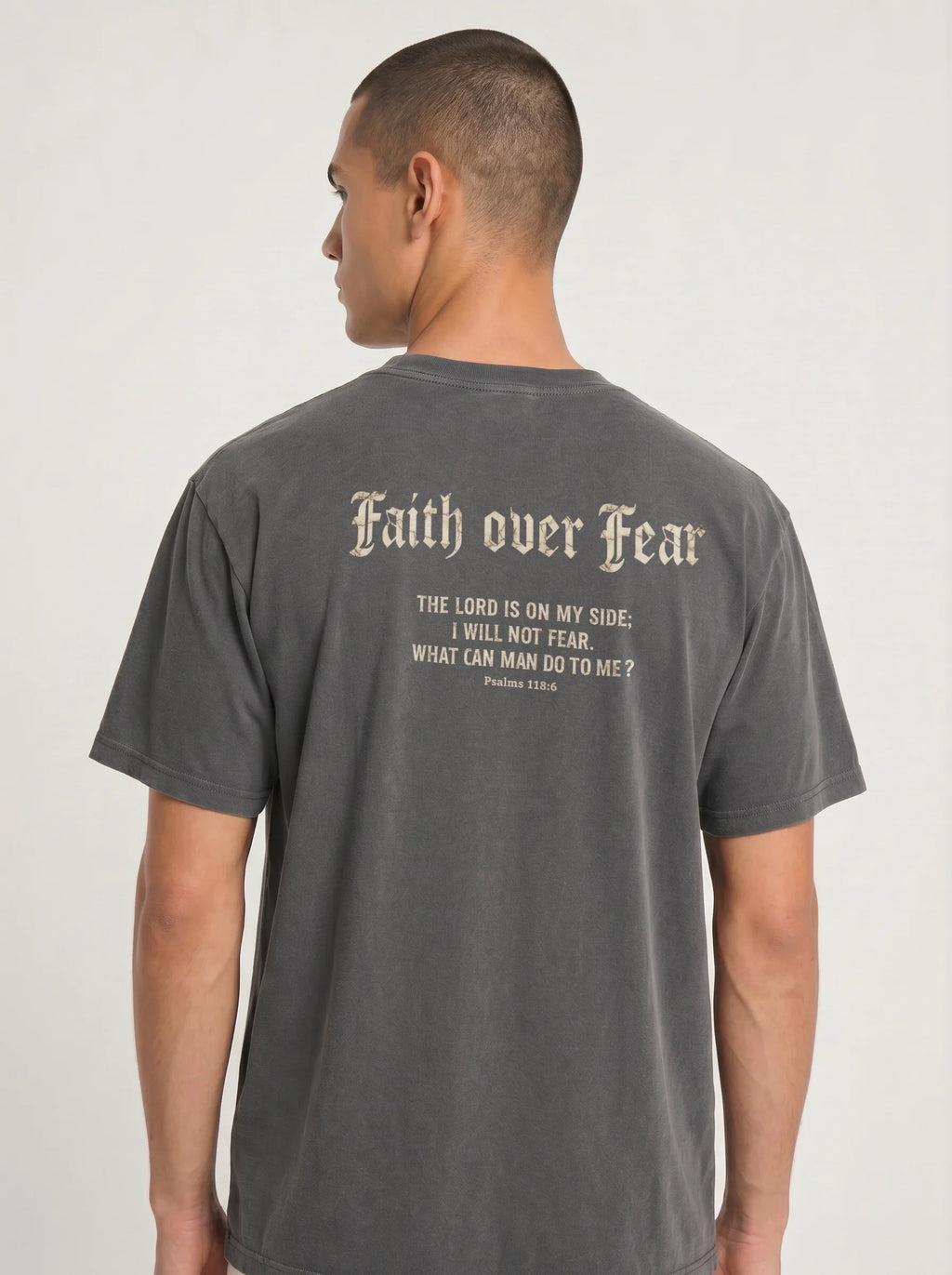 A man viewed from the back wearing a dark gray t-shirt with a distressed cream-colored gothic font graphic that reads 'Faith over Fear' and a bible verse below.