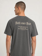 A man viewed from the back wearing a dark gray t-shirt with a distressed cream-colored gothic font graphic that reads 'Faith over Fear' and a bible verse below.