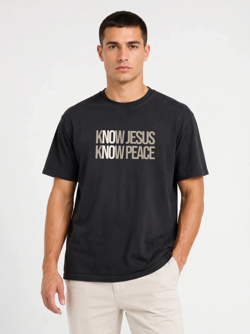 A man wearing a black short-sleeve t-shirt with the text 'KNOW JESUS KNOW PEACE' printed in beige on the chest, paired with light beige pants, against a plain light gray background.