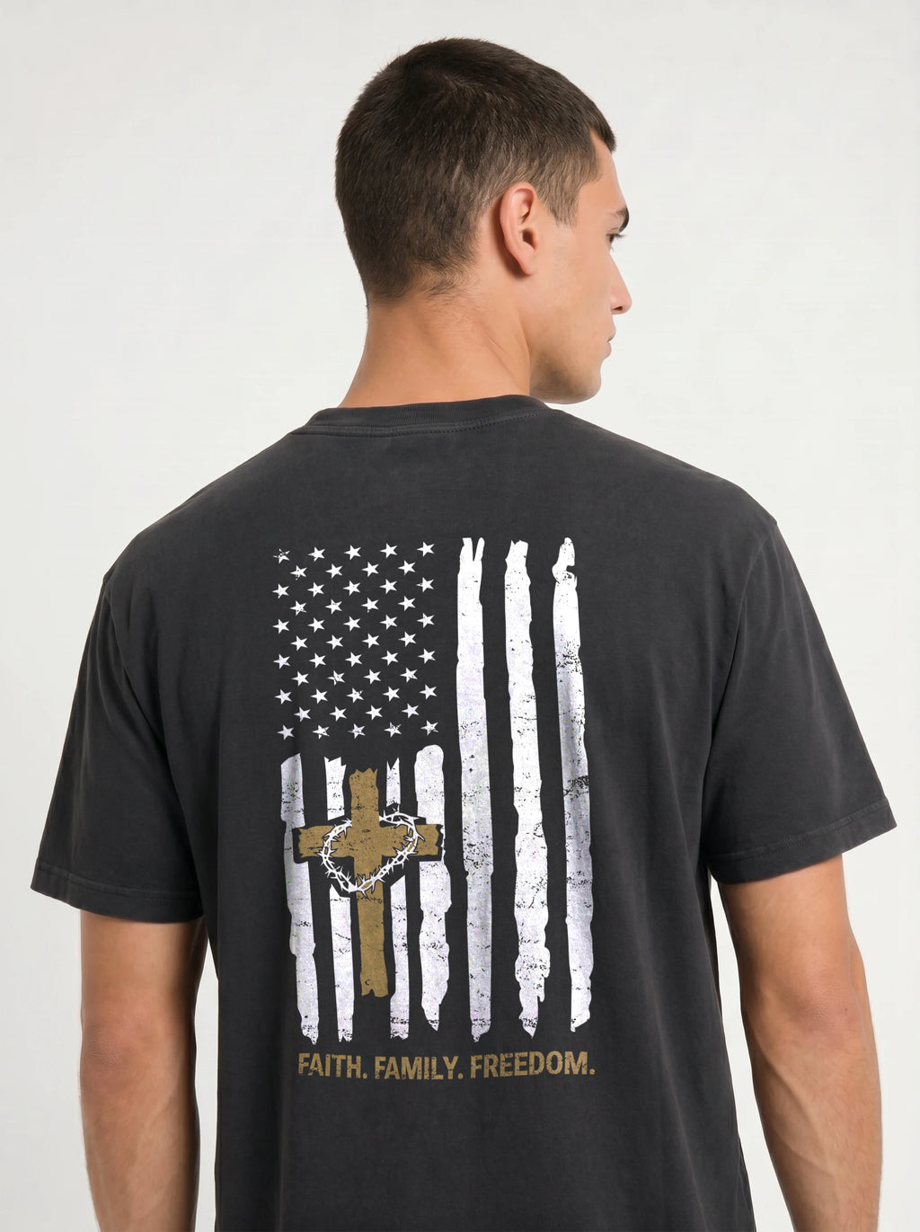 A man wearing a dark charcoal gray t-shirt, viewed from the back. The shirt features a distressed graphic of a vertical American flag with a wooden cross and a hanging rosary in the center, and the words 'FAITH, FAMILY, FREEDOM.'