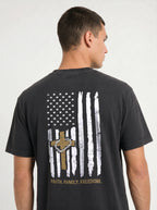 A man wearing a dark charcoal gray t-shirt, viewed from the back. The shirt features a distressed graphic of a vertical American flag with a wooden cross and a hanging rosary in the center, and the words 'FAITH, FAMILY, FREEDOM.'
