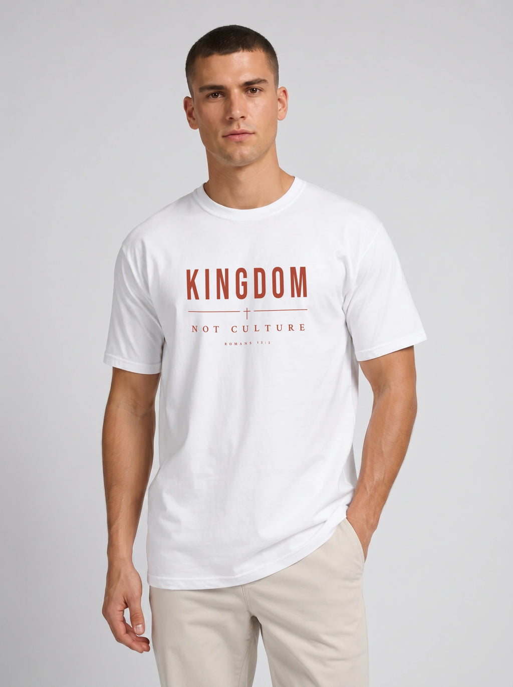 A man wearing a white short-sleeve t-shirt with a red graphic print that reads 'KINGDOM NOT CULTURE' on the chest, paired with beige trousers, against a light gray studio background.
