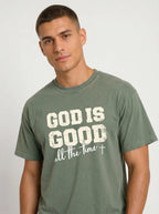 A man wearing a faded olive green t-shirt with a distressed white graphic that reads 'GOD IS GOOD all the time +' on the front, set against a plain light gray background.