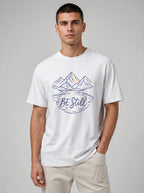 A man wearing a white short-sleeve t-shirt with a graphic of mountains and the text 'BE Still' in navy blue, paired with beige pants, against a neutral gray background.