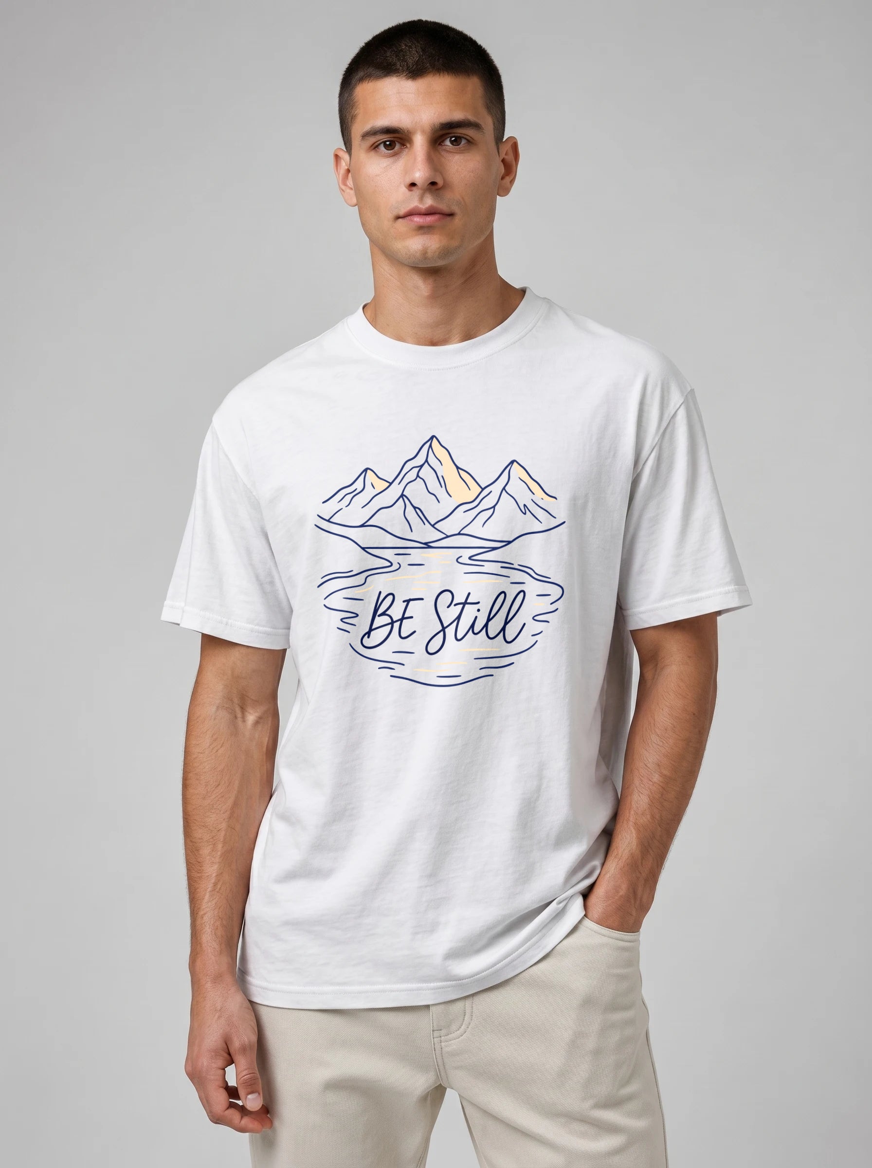 A man wearing a white short-sleeve t-shirt with a graphic of mountains and the text 'BE Still' in navy blue, paired with beige pants, against a neutral gray background.