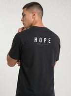 A man wearing a black t-shirt, viewed from the back. The shirt features the word 'HOPE' printed in white block letters with 'ROMANS 15:13' underneath.