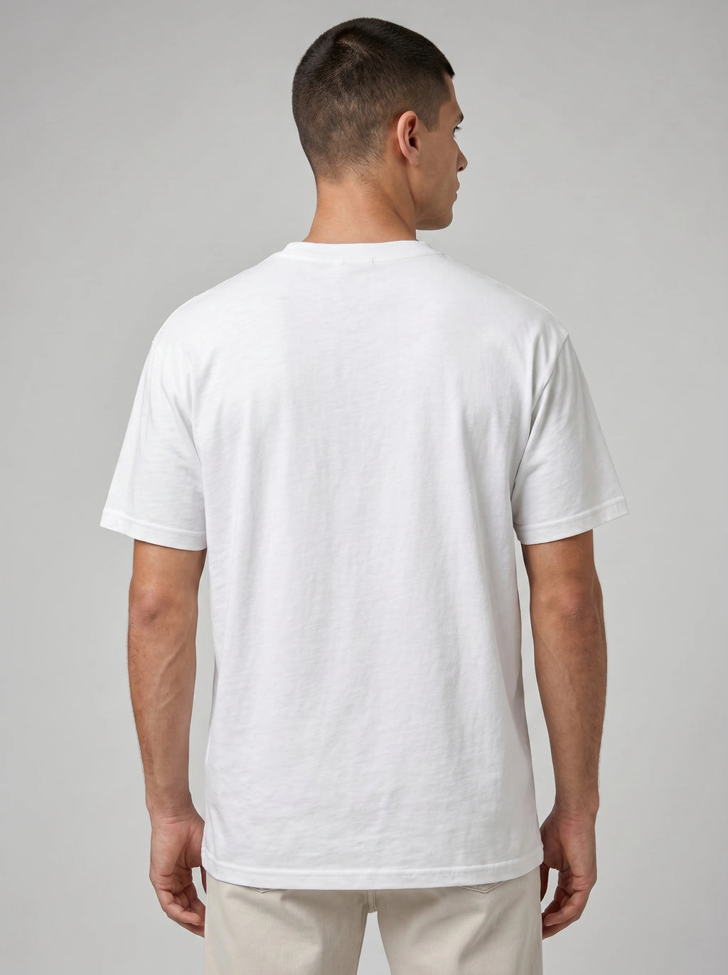 A man viewed from the back wearing a plain white short-sleeve crewneck t-shirt and light beige trousers, set against a neutral gray studio background.