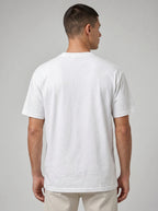 A man viewed from the back wearing a plain white short-sleeve crewneck t-shirt and light beige trousers, set against a neutral gray studio background.