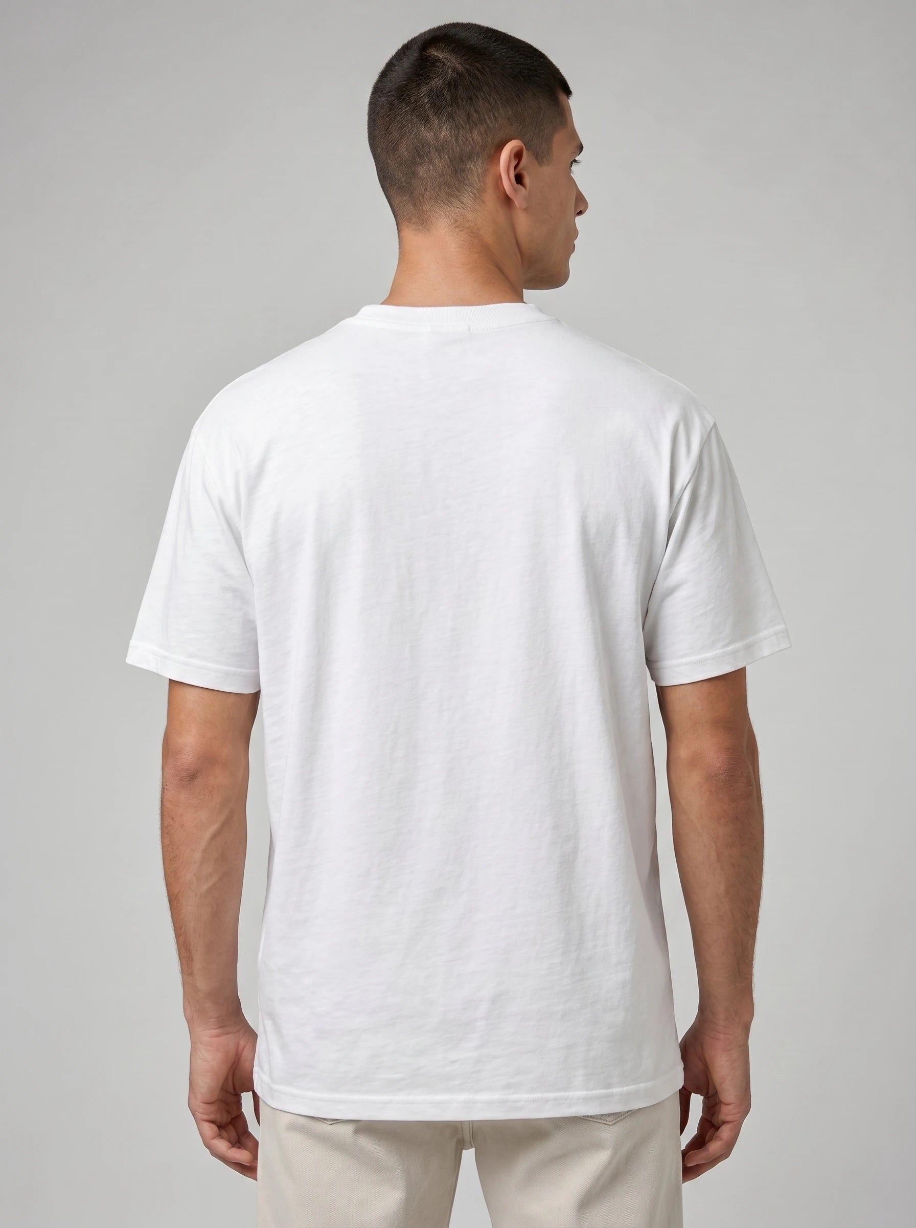 A man viewed from the back wearing a plain white short-sleeve crewneck t-shirt and light beige trousers, set against a neutral gray studio background.