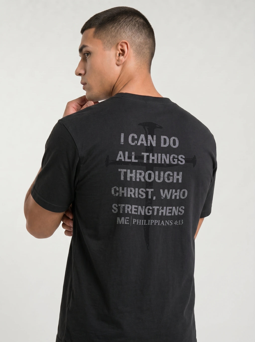 A man wearing a black t-shirt, viewed from the back. The shirt features a distressed gray graphic of a cross and a Bible verse from Philippians 4:13 printed on the back.