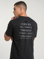 A man wearing a black t-shirt, viewed from the back. The shirt features a distressed gray graphic of a cross and a Bible verse from Philippians 4:13 printed on the back.