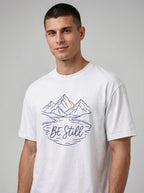 A man wearing a white crewneck t-shirt with a graphic of mountains, a lake reflection, and the text 'Be Still' in blue and yellow, set against a plain gray studio background.