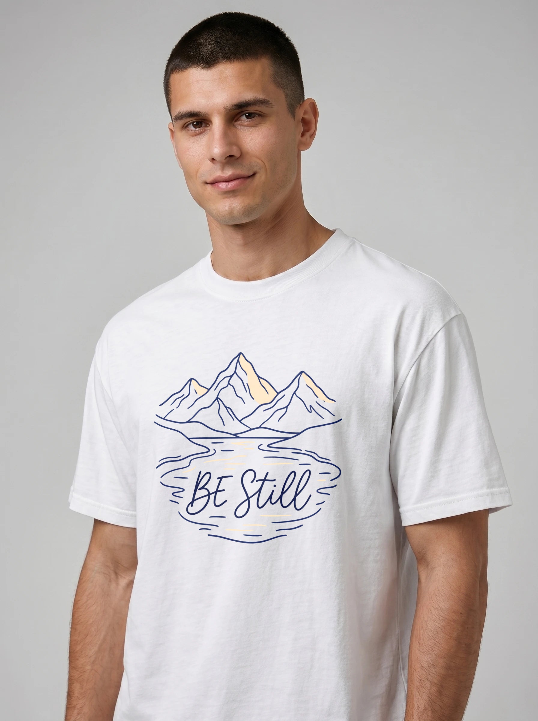 A man wearing a white crewneck t-shirt with a graphic of mountains, a lake reflection, and the text 'Be Still' in blue and yellow, set against a plain gray studio background.