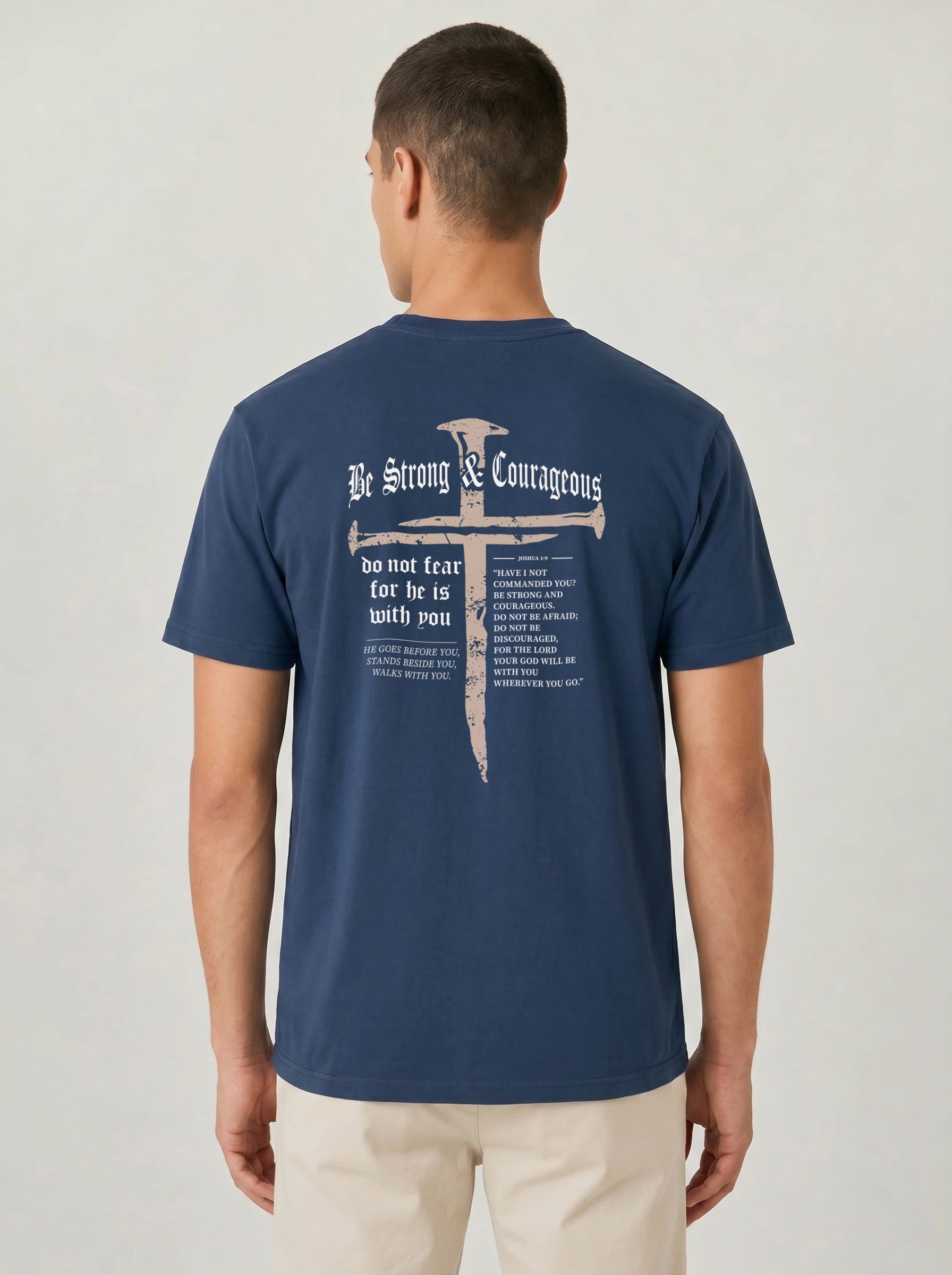 A man viewed from the back wearing a navy blue t-shirt with a large white graphic print on the back featuring a cross,  and the text 'Be Strong & Courageous'.