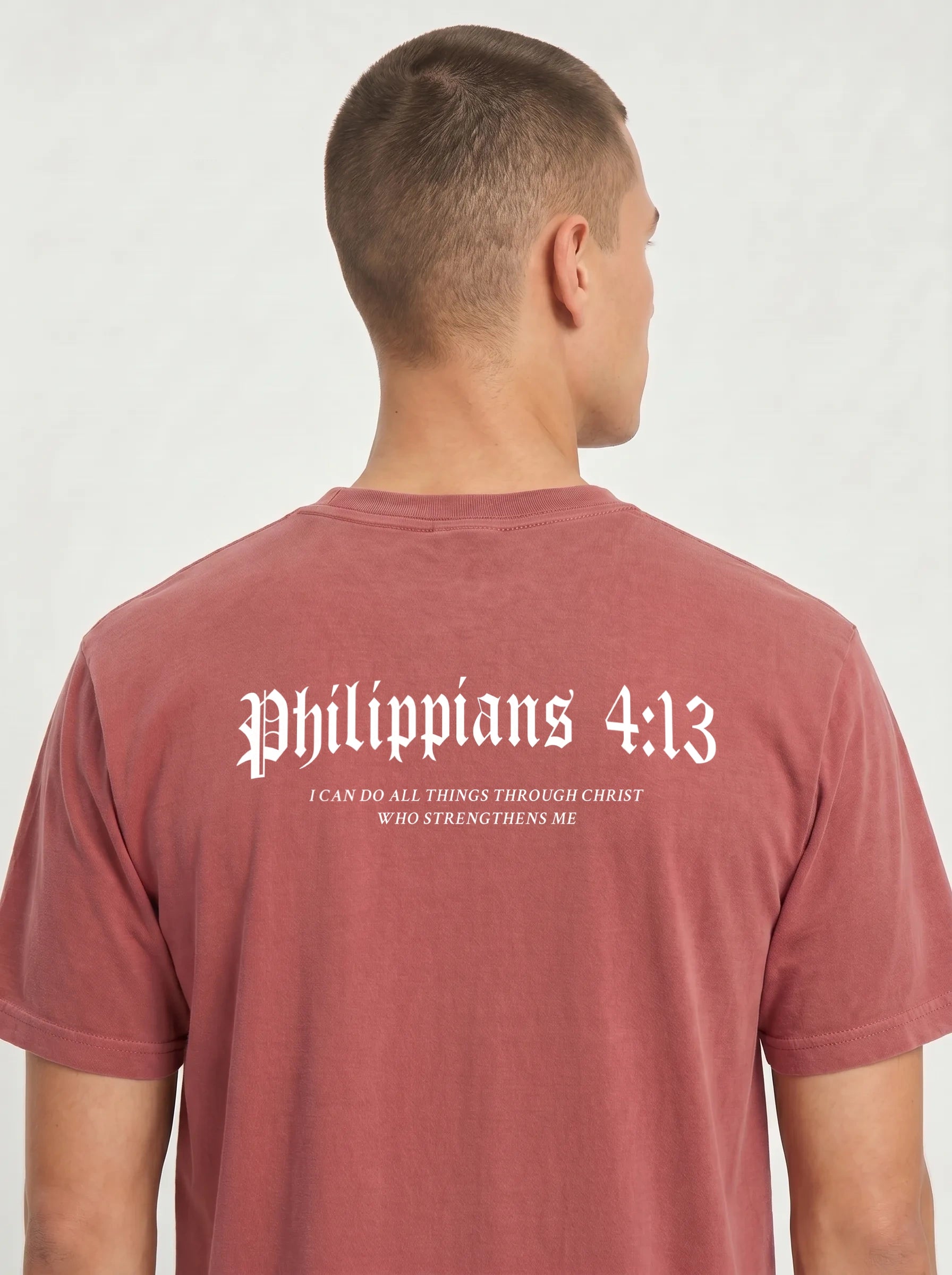 A man viewed from behind wearing a dusty rose short-sleeve t-shirt with white Gothic-style text reading 'Philippians 4:13' and smaller scripture text below, against a light gray background.