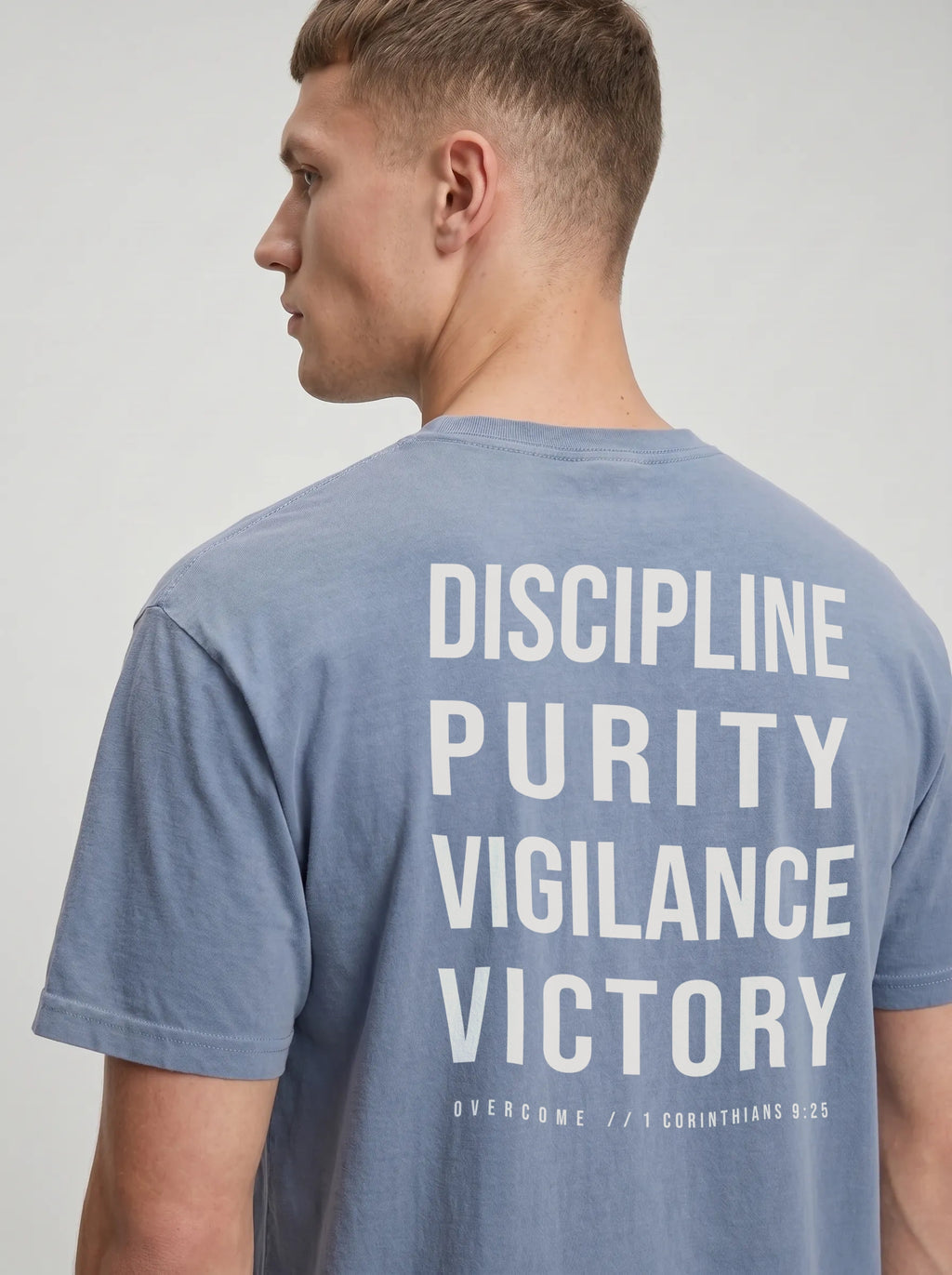 Person wearing a faded blue T-shirt with large back text reading “Discipline Purity Vigilance Victory” and “Overcome // 1 Corinthians 9:25.”