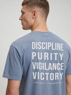 Person wearing a faded blue T-shirt with large back text reading “Discipline Purity Vigilance Victory” and “Overcome // 1 Corinthians 9:25.”