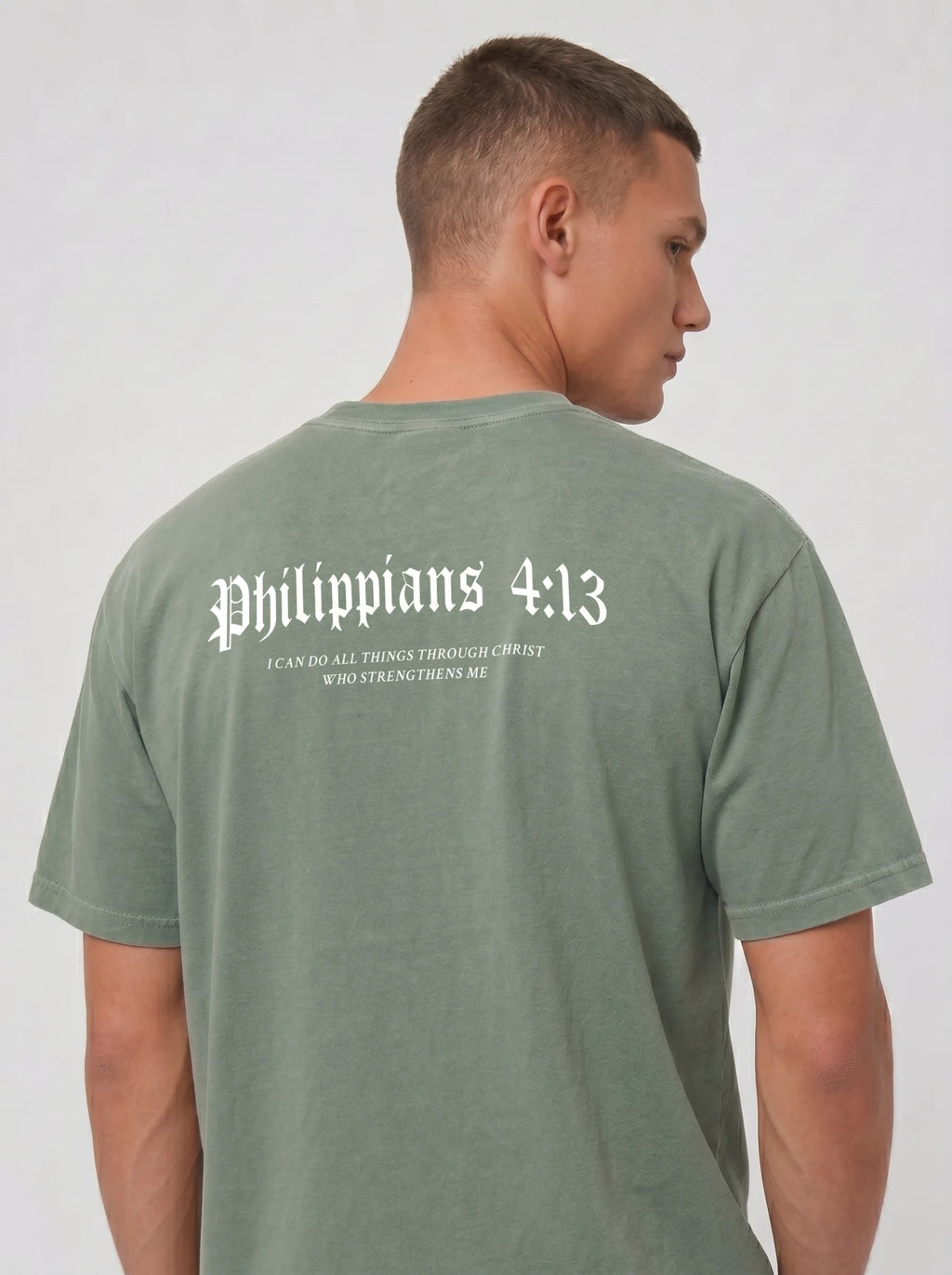 A man wearing an olive green t-shirt, viewed from the back. The shirt features the text 'Philippians 4:13' in a white Gothic font, with smaller white text underneath reading 'I CAN DO ALL THINGS THROUGH CHRIST WHO STRENGTHENS ME'.