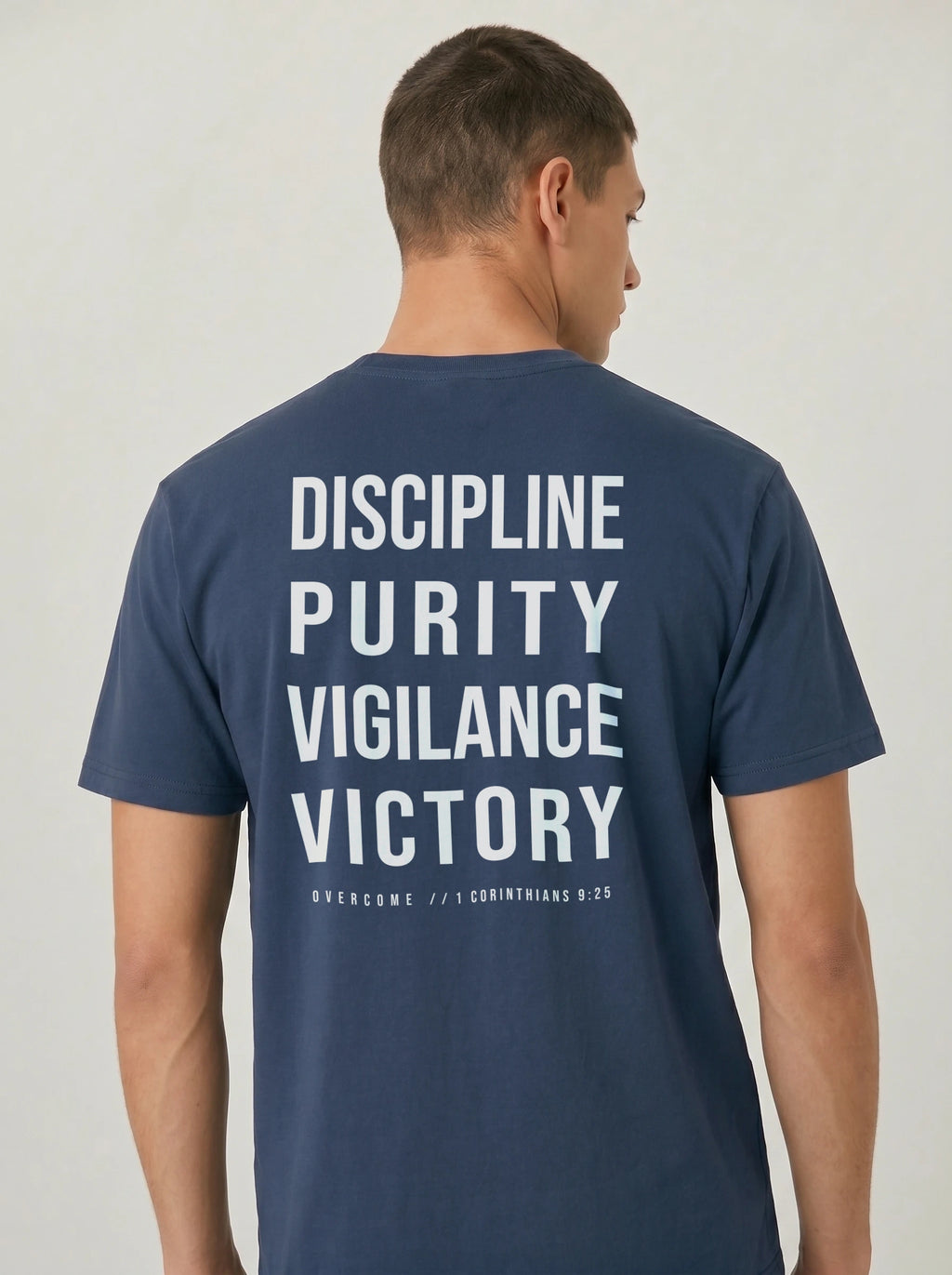 Person wearing a navy T-shirt with large back text reading “Discipline Purity Vigilance Victory” and “Overcome // 1 Corinthians 9:25.”