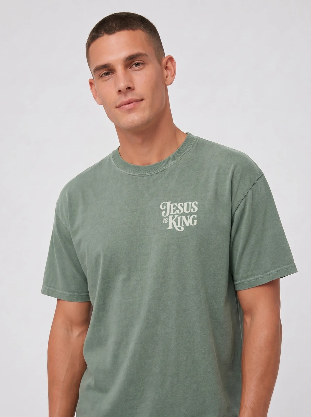 Person wearing a faded green T-shirt with a small cream “Jesus is King” design on the left chest.
