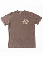 Brown T-shirt laid flat with a small white and orange chest graphic reading “God Over All Things.”