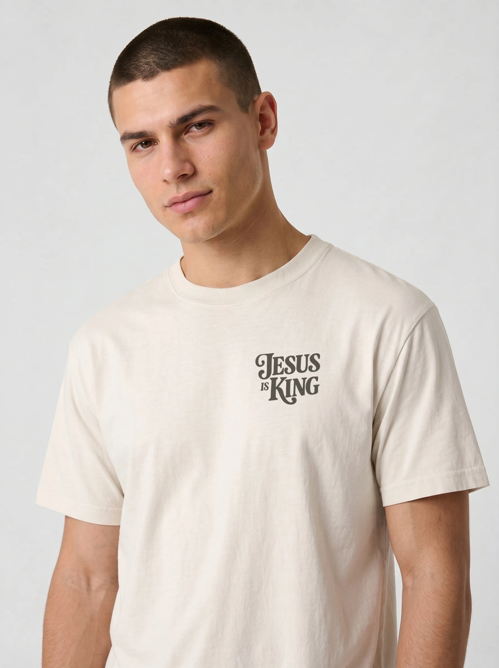 Person wearing a cream T-shirt with a small dark “Jesus is King” design on the left chest.