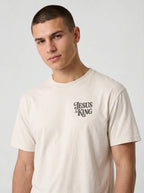 Person wearing a cream T-shirt with a small dark “Jesus is King” design on the left chest.