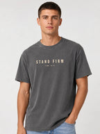 Person wearing a faded gray T-shirt with distressed beige text reading “Stand Firm” and “1 Cor. 16:13.”