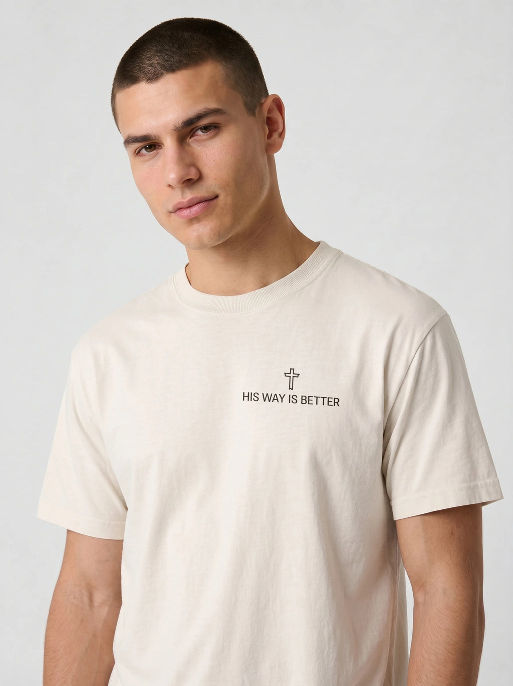 A man wearing a light beige crewneck t-shirt with a small black graphic on the chest featuring a cross and the text 'HIS WAY IS BETTER'.