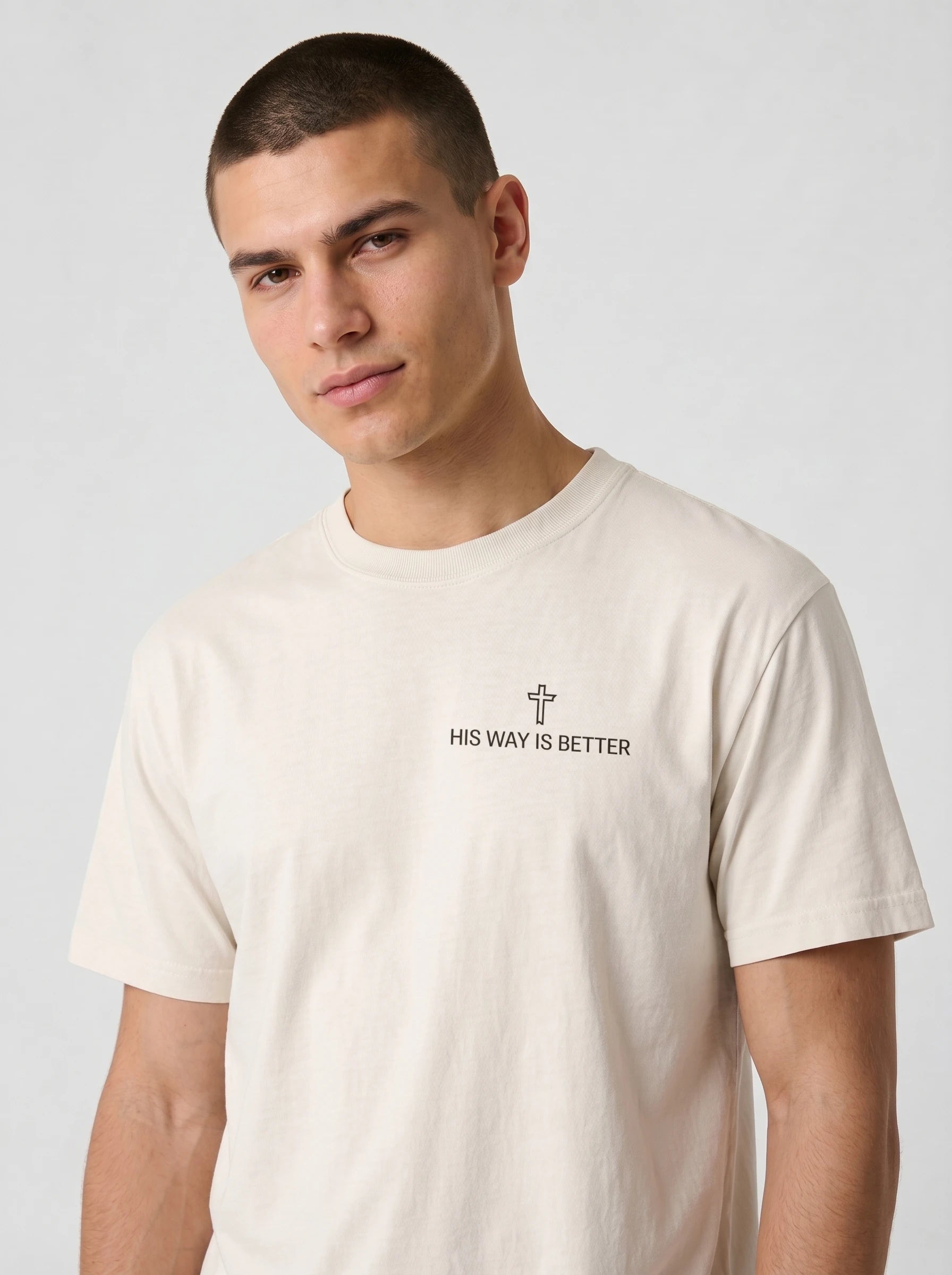 A man wearing a light beige crewneck t-shirt with a small black graphic on the chest featuring a cross and the text 'HIS WAY IS BETTER'.